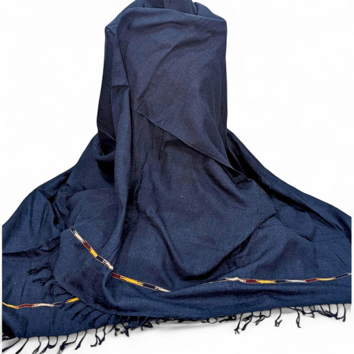 A blue jeans, subtly textured shawl with a thin, patterned border and a delicate fringed edge is draped and partially folded against a plain white background.