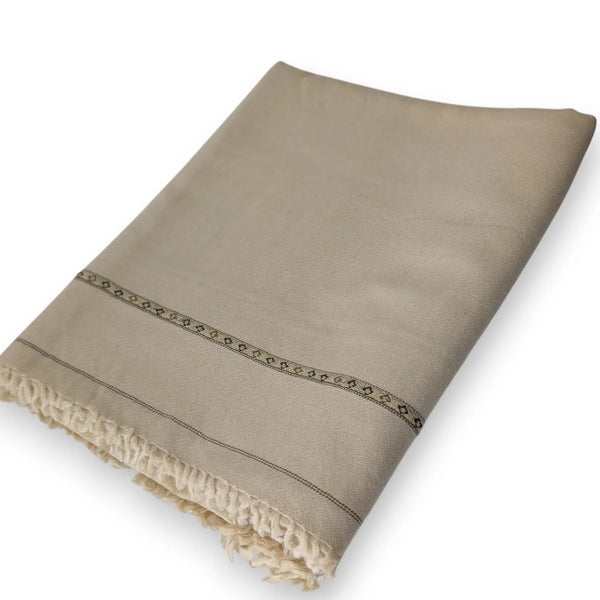 A light tan, subtly textured shawl with a thin, patterned border and a delicate fringed edge is draped and partially folded against a plain white background.