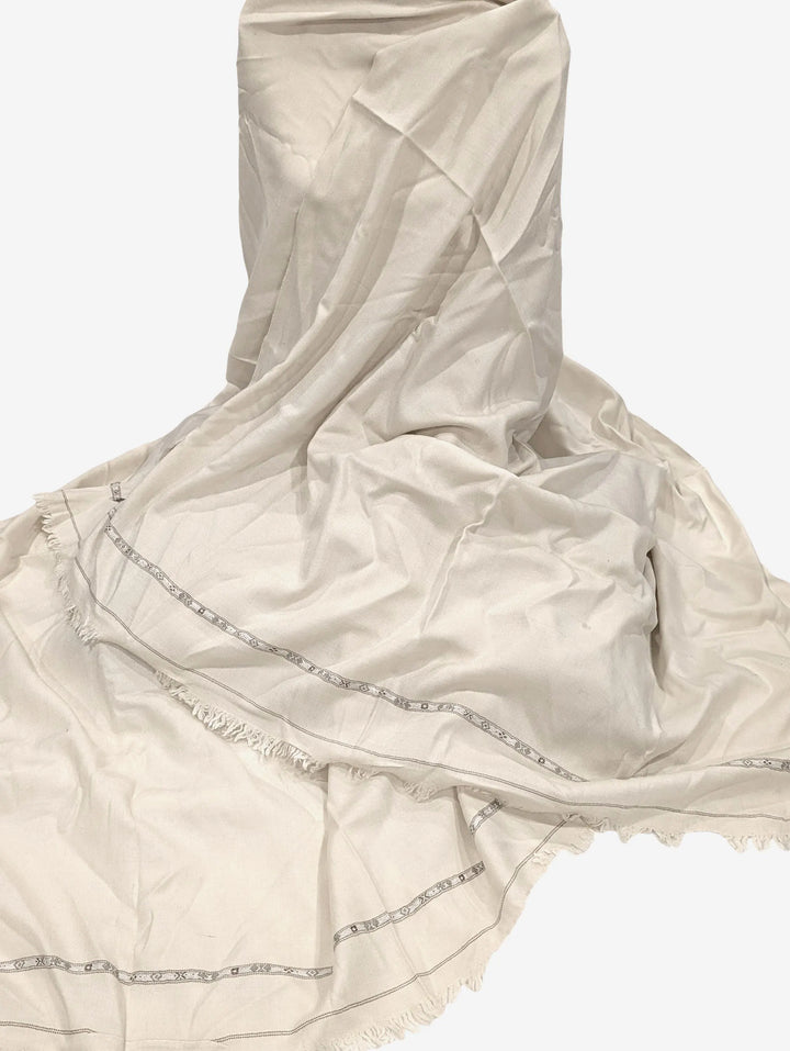 An off-white, subtly textured shawl with a thin, patterned border and a delicate fringed edge is draped and partially folded against a plain white background.